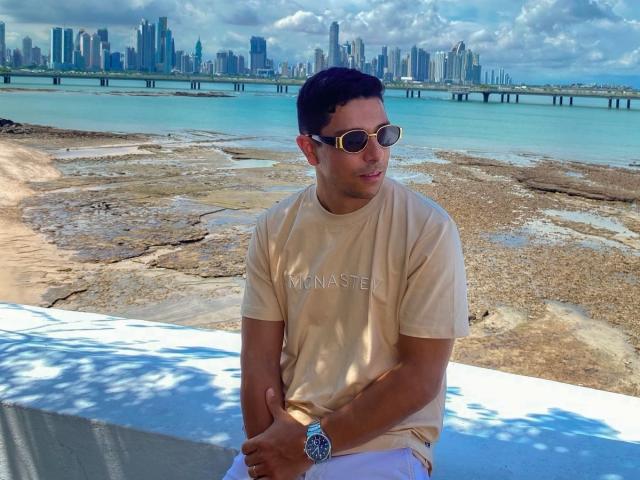 JoseLuiss with short dark hair, wearing round sunglasses, beige t-shirt, white pants, and a watch, sitting on a blue-white surface, looking at the camera.