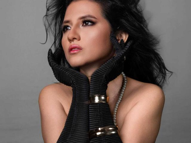 RenataRivas with black ribbed gloves, holding her face, looking up with dark eyes, wearing silver bracelets, and a pearl necklace, against a gray background.