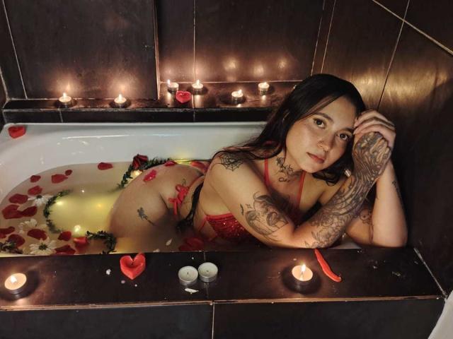 Gaby Mathew trägt ein rotes Spitzen-Dessous-Set, hat Tattoos an den Armen und befindet sich in einer dunklen, mit Fliesen ausgekleideten Badewanne mit Kerzen, Rosenblättern und Wasser. Ihr Hintern ist angehoben, und sie schaut in die Kamera..