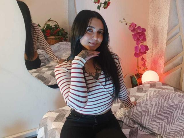 GabyMathew with long black hair, striped shirt, black pants, tattoo on chest, sitting on bed, smiling, hand on chin, pink orchid, round mirror, striped pillow, white wall.