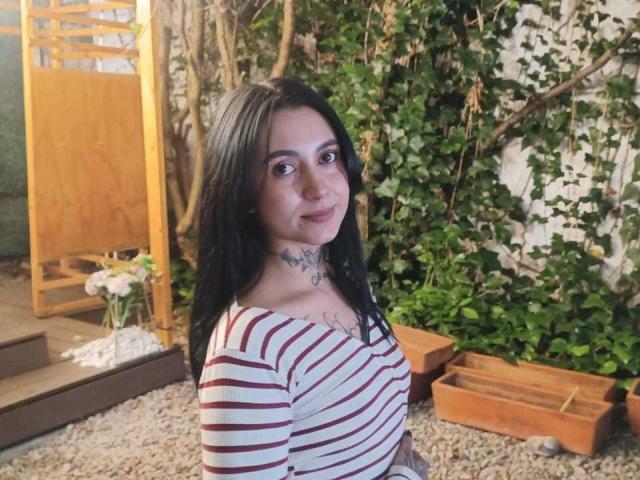 GabyMathew with black hair, striped shirt, tattoos, and a slight smile, standing in a gravel yard with green plants and wooden panels. Her tits are small and she's cute.