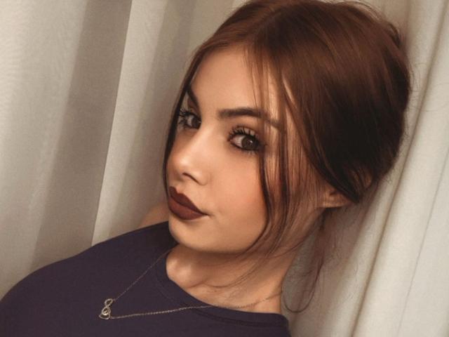 SweetGiggles69 with brown hair in a bun, dark lipstick, and a navy top, wearing a necklace, has a cute, slightly smug expression, looking at the camera.