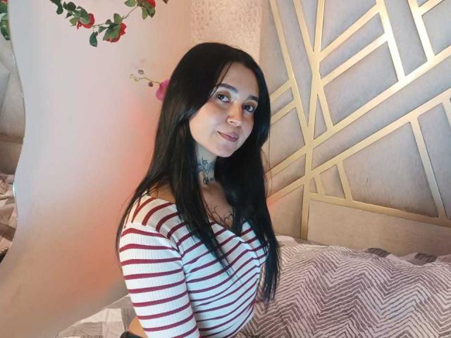 GabyMathew with long black hair, wearing a red and white striped top, showing her tits, has a tattoo on her neck, and a nose ring, sitting on a bed.