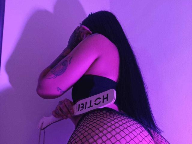 GabyMathew with long black hair, tattooed arm, wearing fishnet pants, black strap top, holding "BLICH" sign, purple-lit, posing with back to camera, showing her ass.