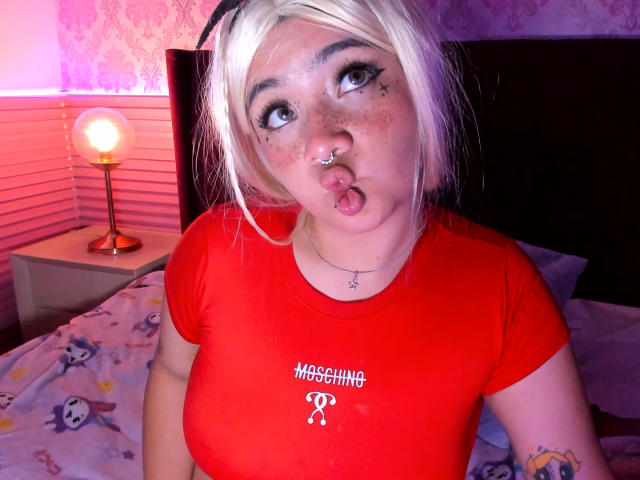 AmaraKazumi with blonde hair, nose ring, and freckles, pouting with her tongue out, wearing a red shirt, showing her tits, in a bedroom with a lamp.
