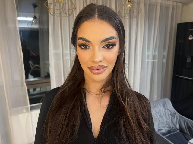 KendallJollie has long dark brown hair, thick eyebrows, and is sticking her tongue out slightly. She's wearing a black top with a necklace, looking sexy.