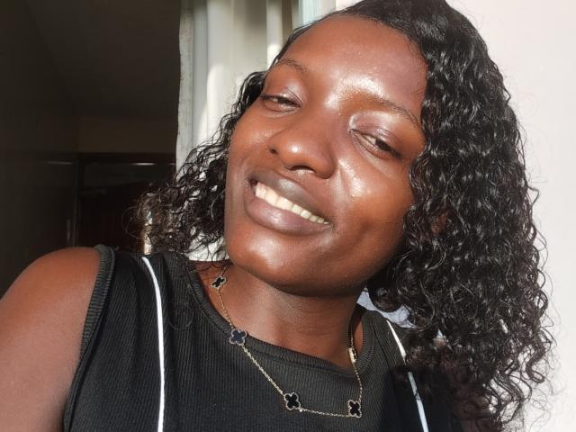 SweetCrimson has dark brown skin, curly black hair, and is smiling with eyes closed. She's wearing a black sleeveless top and a necklace with small black stars.