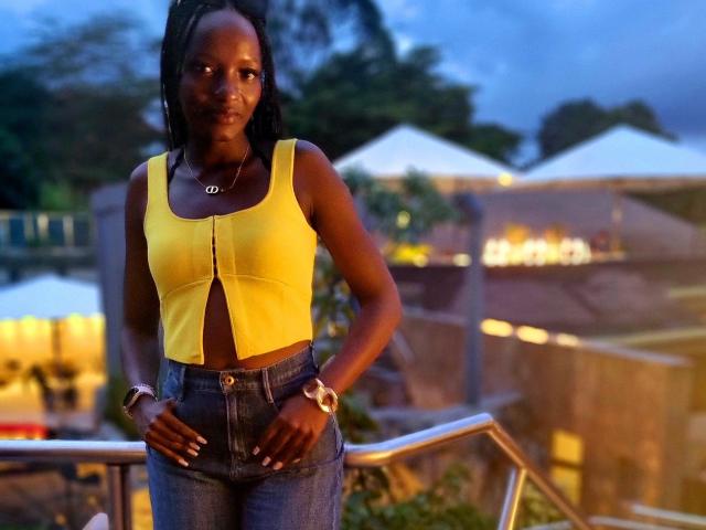 SweetCrimson with dark skin, wearing a yellow sleeveless top showing her small tits, and blue jeans, standing with hands on hips, blue evening sky in background.