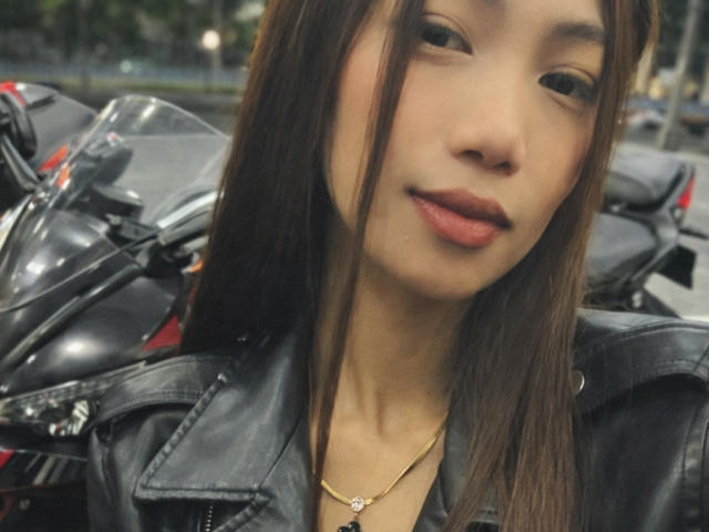 SweetPinkieFrench with straight brown hair, wearing a black leather jacket, has a small gold necklace with a diamond pendant, and a slight smile, standing next to a motorcycle.