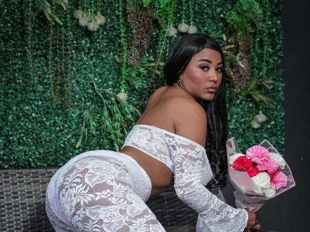 Grettchen with dark skin, long black hair, in white lace off-shoulder top and matching high-waist pants, holding pink and white flowers, bending over, showcasing her big ass.