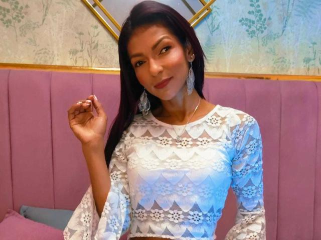 HannaPretty with long dark hair, wearing a white lace dress, holding a ring, pink upholstered background, and large silver earrings, smiling slightly. Boobs visible under lace pattern.