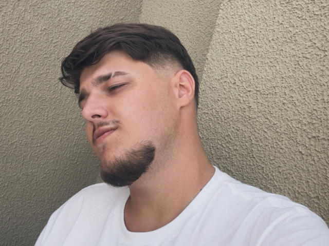 BigJota has a medium skin tone, short brown hair, and a stubby beard, wearing a white shirt, with a relaxed, slightly smug expression, eyes half-closed.