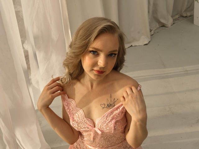 EmilySunshineS with blonde hair, pink lace lingerie, holding straps, showing small tits, tattoo on chest, looking up, smiling, white curtains, soft lighting, pale skin.