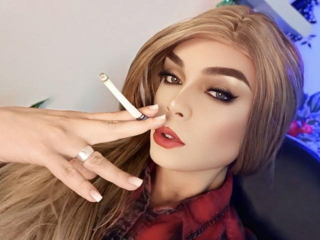 KarolElegance with blonde hair, red lipstick, and dark eyeliner, holding a cigarette to her lips. Her hand has a silver ring and French-manicured nails.