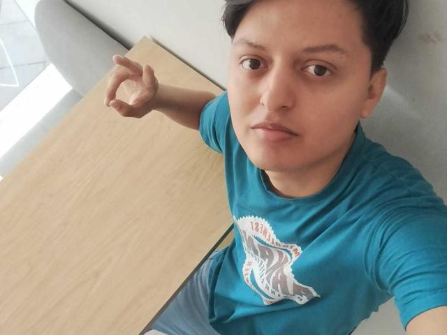 KevinCharles with short black hair, brown eyes, wearing a teal shirt with a white graphic, standing by a wooden table, making a peace sign with his right hand.