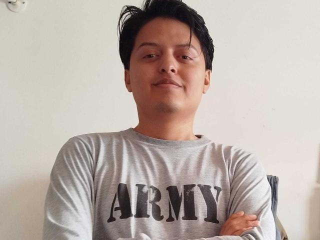 KevinCharles, medium-build Asian man with short black hair, slight smile, wearing a grey "ARMY" shirt, arms crossed, standing against a plain white wall.