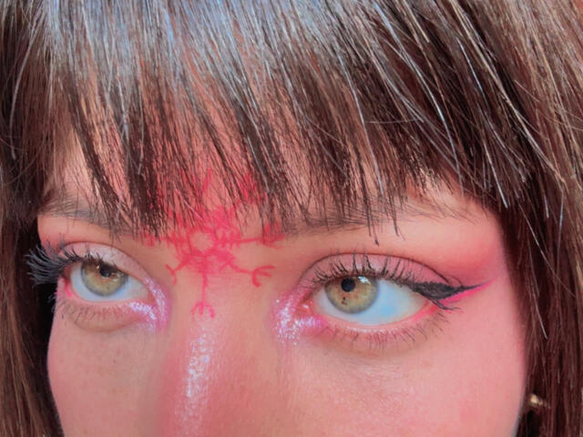 IzzysMisss's face with brown bangs, pink eyeshadow, and red henna pattern on forehead, blue eyes, and black eyeliner. Boobs not visible, focus on eyes and forehead.