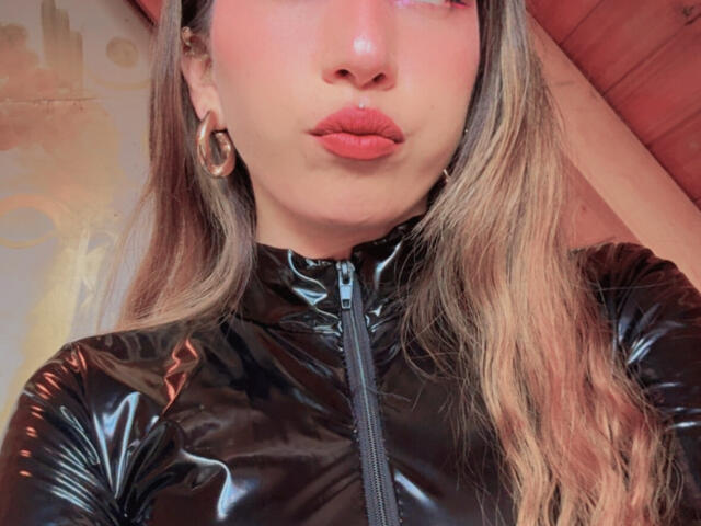 IzzysMisss with pouty lips, wearing a shiny black latex zip-up, gold hoop earrings, and long blonde hair, has a small piercing below her nose.