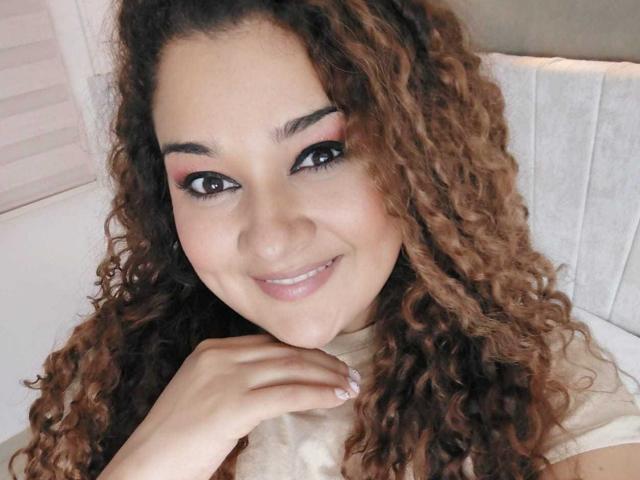 NataliaWendys with curly brown hair, pink eye makeup, and a smile, holding her chin, showing her small nose and cute face, wearing a beige top.
