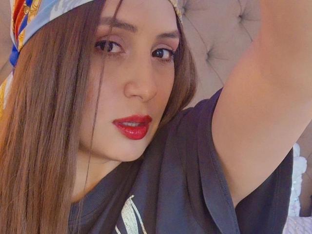 IsabelSoler with long brown hair, red lipstick, black shirt, and colorful cap, raising her arm in a selfie, showing off her sexy face and cute smile.