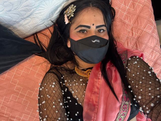DaliaDubey lying on pink quilted bed, wearing black fishnet top with pearl details, black mask, pink scarf, gold necklace, black hair with pink hair accessory, dark makeup.