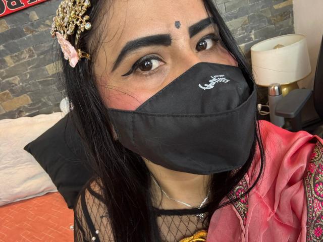 Dalia Dubey, con cabello oscuro, ojos marrones, máscara negra, tocado ornamentado, ropa tradicional rosa y roja, nariz y parte superior del pecho visibles, top de malla negra, fondo de pared de piedra..