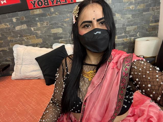DaliaDubey with long black hair, wearing a black mask, sheer fishnet top, pink and red traditional outfit, and a gold necklace, sitting on an orange bed.