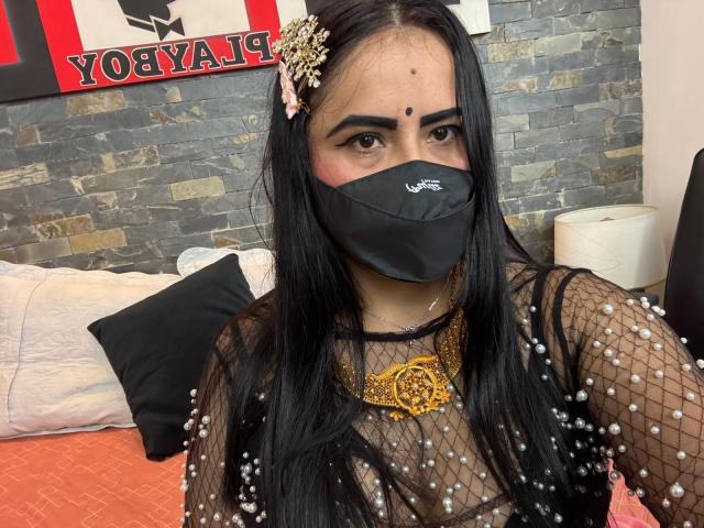 DaliaDubey with long black hair, wearing a black mask, sheer fishnet top with pearl details, gold necklace, and pink hairpin, sitting on an orange bed.