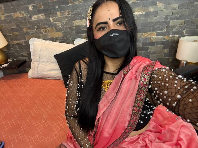 DaliaDubey with long black hair, wearing a pink sari, black mask, and beaded top, sitting on a bed, showing her tits, with a stone wall background.