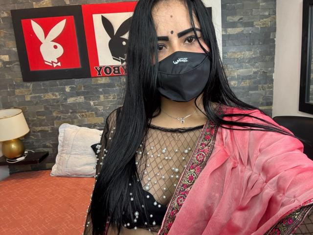 DaliaDubey with long black hair, wearing a black mask, sheer top with beads, pink robe, and a black bra, showing her tits. She has a small nose dot.
