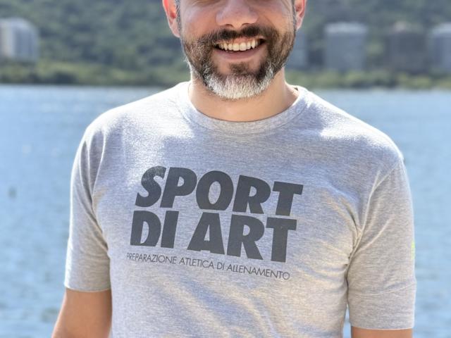 GrisalhoRoludo has a gray beard and is smiling, wearing a gray sport di art t-shirt, showing his upper body, with a blue water background.