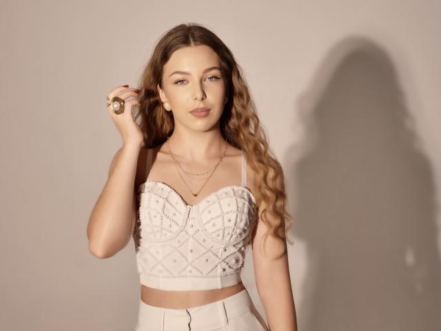 LilyBlake with long wavy brown hair, wearing a white beaded strapless top, gold ring, and necklace, standing against a plain background with a shadow on the right.