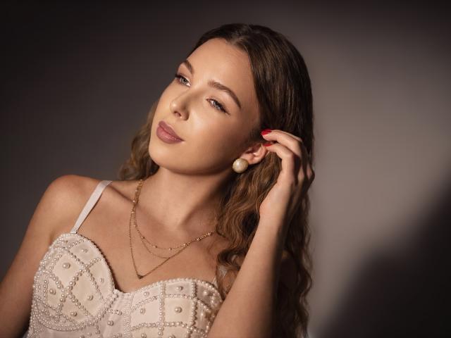 LilyBlake with wavy brown hair, wearing a beaded white dress, holding a pearl earring, looking slightly up with a subtle smile, and red-painted nails.