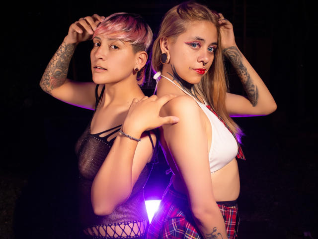 Two women stand back-to-back, arms raised. HadeDanny on the left has short pink hair, black mesh top, tattoos on arms, and ear gauges. HadeDanny on the right has long blonde hair, white crop top, red plaid skirt, tattoos on arms, and black ear gauges. Both have red lipstick. A purple light source illuminates from behind. The image is sharp with vivid colors. The composition emphasizes their tattoos and contrasting hair styles.