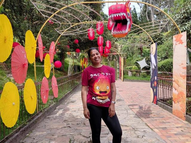 PauletteLoveX stands smiling, wearing a red shirt with a fire face, black pants, and a wristwatch, under a yellow and red lantern arch with a hanging red mouth.