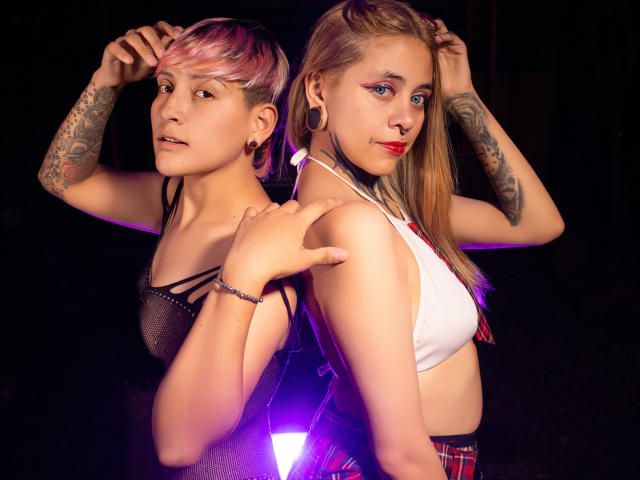 Two HadeDaniels with tattoos, back-to-back, one with pink hair in mesh top, other with blonde hair in white top, red plaid skirt, purple light, both looking at camera.