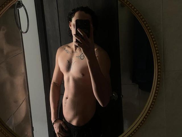 JayTaylor torse nu, avec des tatouages, tenant un téléphone, autoportrait devant un miroir, cheveux foncés, silhouette mince, main dans le pantalon, peau bronzée, tatouage visible sur la poitrine, petits tétons, porte sombre, miroir rond..