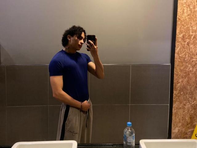 JayTaylor, un homme musclé avec des cheveux noirs bouclés, porte une chemise bleue moulante et un pantalon gris, et prend un selfie devant un miroir dans une salle de bain, sa main droite tenant son téléphone. Bouteille d'eau..
