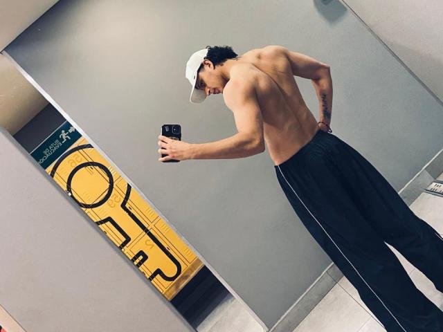 JayTaylor, torse nu avec un dos musclé et un pantalon noir, prend un selfie devant un miroir dans une salle de bain, portant une casquette blanche et tenant un téléphone noir. Ses fesses sont visibles..