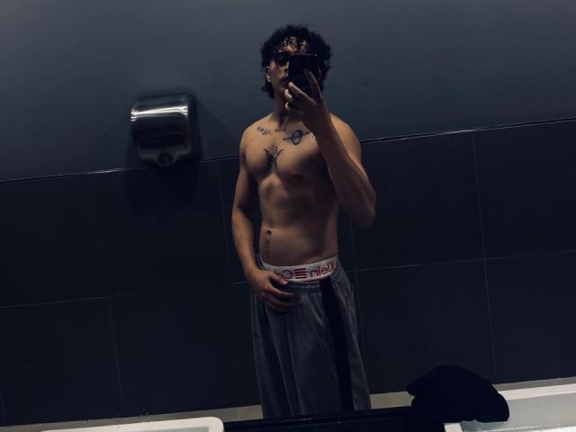 JayTaylor sans chemise, avec des cheveux noirs bouclés, des tatouages sur la poitrine, tenant un téléphone, portant un pantalon gris, debout dans une salle de bain faiblement éclairée, une main couvrant l'aine, reflet dans un miroir, mur noir, papier toilette..