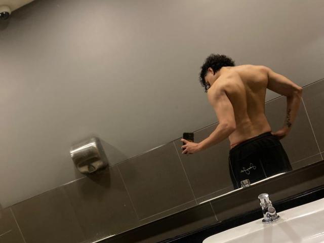 Jay Taylor mit einem trainierten, nackten Oberkörper, schwarzen, lockigen Haaren und schwarzen Hosen, der in einem Badezimmer in einen Spiegel schaut, ein Telefon in der Hand hält, unter ihm ein Waschbecken und ein Wasserhahn..