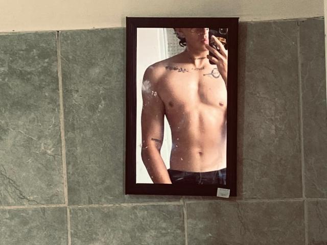 JayTaylor sans chemise, avec des tatouages, tenant un téléphone, reflet dans un miroir montrant des abdominaux, une poitrine et un jean foncé, mur en arrière-plan avec des carreaux verts, léger effet de flou sur le reflet du miroir..