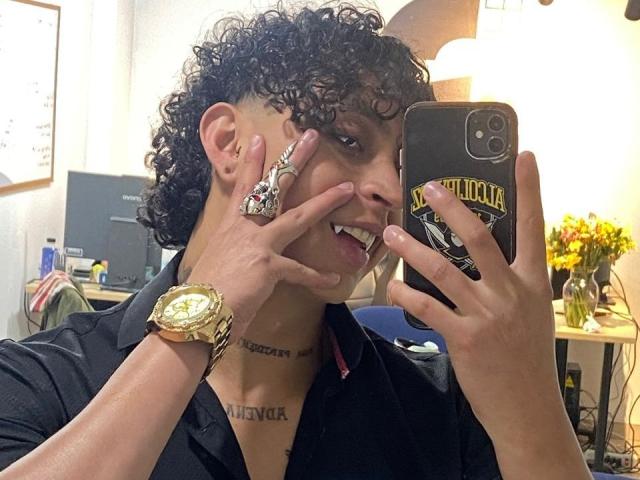 Jay Taylor, con i capelli neri ricci, tiene in mano un telefono con una custodia nera, indossa un orologio d'oro, un anello d'argento, una camicia nera e mostra un tatuaggio, sorridendo..