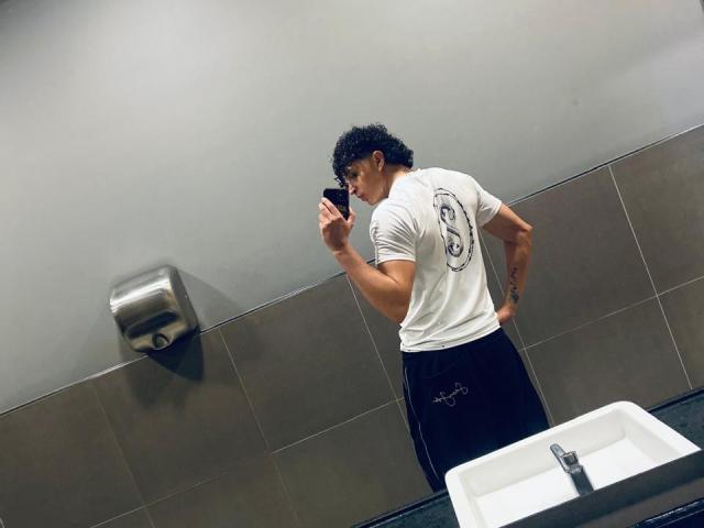 JayTaylor, avec ses cheveux noirs bouclés et ses lunettes, se tient dans une salle de bain, vêtu d'un t-shirt blanc et d'un pantalon noir, et regarde son téléphone, sa main droite posée sur sa hanche..