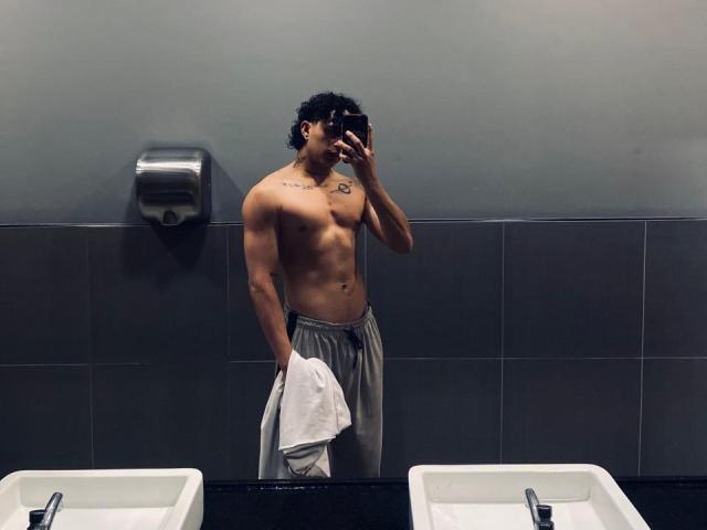 JayTaylor est un homme torse nu, avec une musculature tonique, tenant une serviette blanche d'une main, portant un pantalon gris, debout dans une salle de bain, et prenant un selfie devant un miroir..