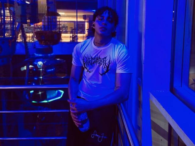 Jay Taylor mit lockigem Haar, in blauem Licht, trägt ein weißes T-Shirt mit Grafik, schwarze Hose, hält eine Flasche, steht neben einem Fenster, blickt leicht nach oben, Hände in der Nähe der Hüfte, entspannt..