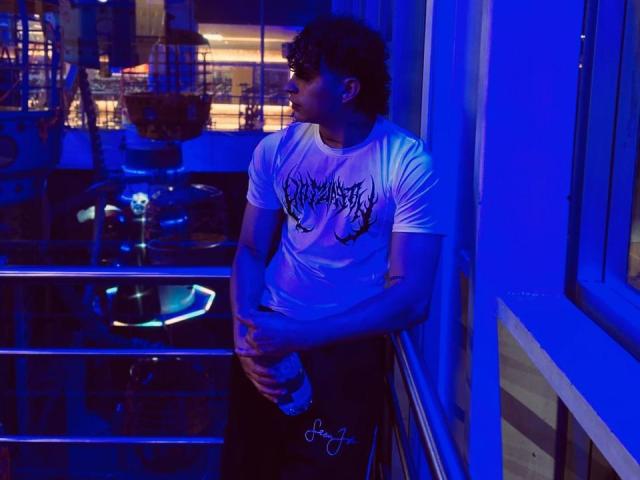 Jay Taylor, com cabelo cacheado, vestindo uma camiseta branca com estampa e calças pretas, está em pé sob uma luz azul neon, segurando uma garrafa, apoiado em um corrimão, olhando para o lado..