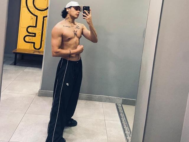 JayTaylor ist ein muskulöser Mann ohne Hemd, mit einer weißen Mütze, schwarzen Hosen und Tattoos auf der Brust, der ein Selfie vor einem Spiegel im Fitnessstudio macht und dabei seine Bauchmuskeln zur Schau stellt..