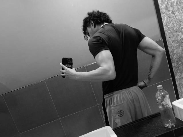 JayTaylor, musclé et avec des cheveux bouclés, porte un t-shirt moulant et un pantalon ample, et prend un selfie dans une salle de bain, montrant ses fesses et un tatouage sur sa main droite..