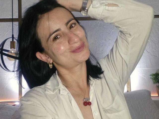 BrendaCantorel has black hair, light skin, small tits, and a cute smile. She's wearing a white shirt, red cherry necklace, and gold earrings, with one arm raised.